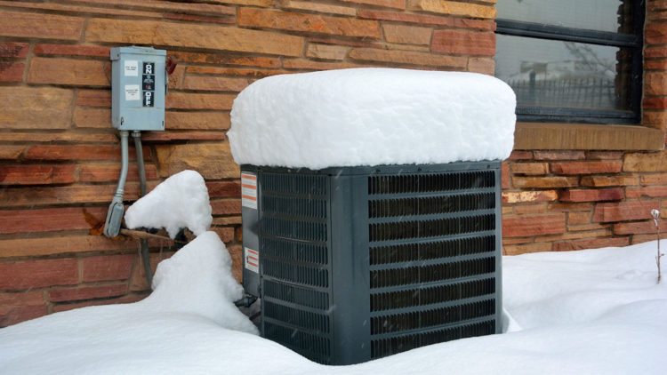 A snow covered air conditioner in front of a brick wall.