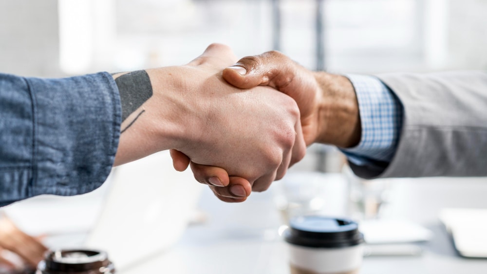 Two individuals shaking hands over a table with coffee cups and a laptop in view.