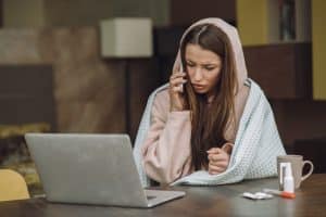A person wearing a hoodie and blanket talks on the phone while sitting at a table with a laptop, medicine, and mug.