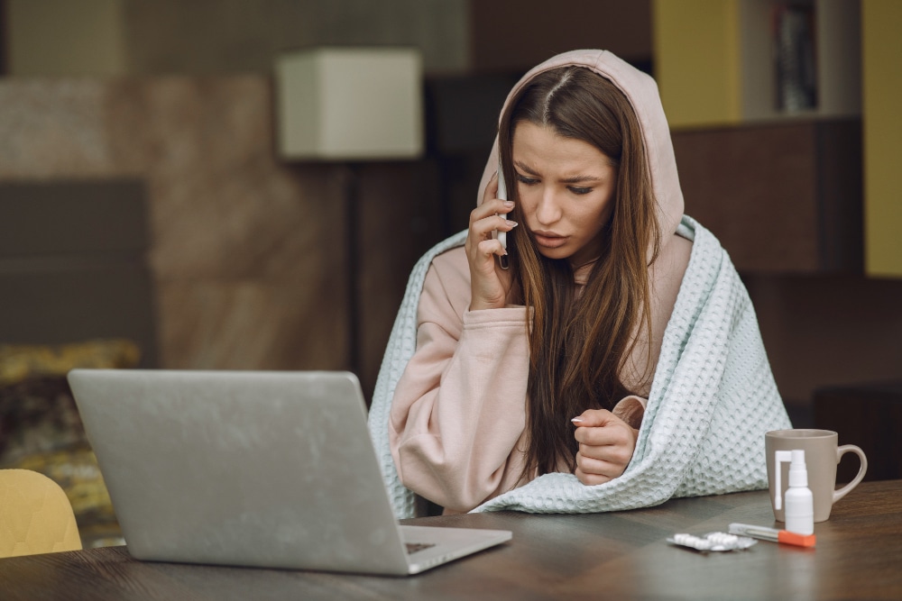 A person wearing a hoodie and blanket talks on the phone while sitting at a table with a laptop, medicine, and mug.