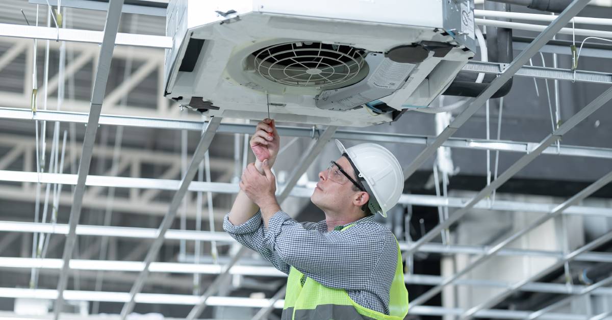 A worker in a hard hat and safety vest repairs a ceiling-mounted air conditioning unit using a screwdriver.